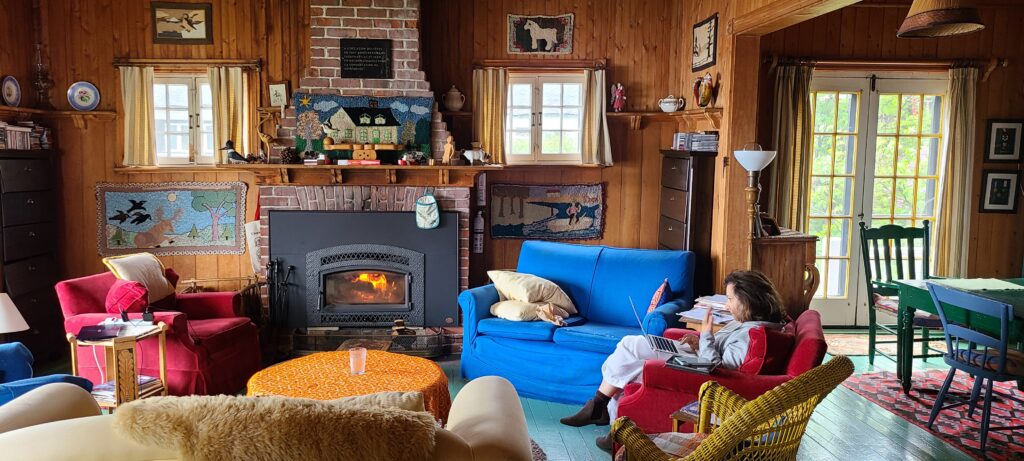 A photo of the inside of Fletcher Cottage during the day, which includes two soft armchairs on either side of a sofa and next to an active fireplace. Maryse Warda sits in a  chair, focused on a laptop. Behind her to the left of the image is a dining room table and french doors leading to the forest outdoors.